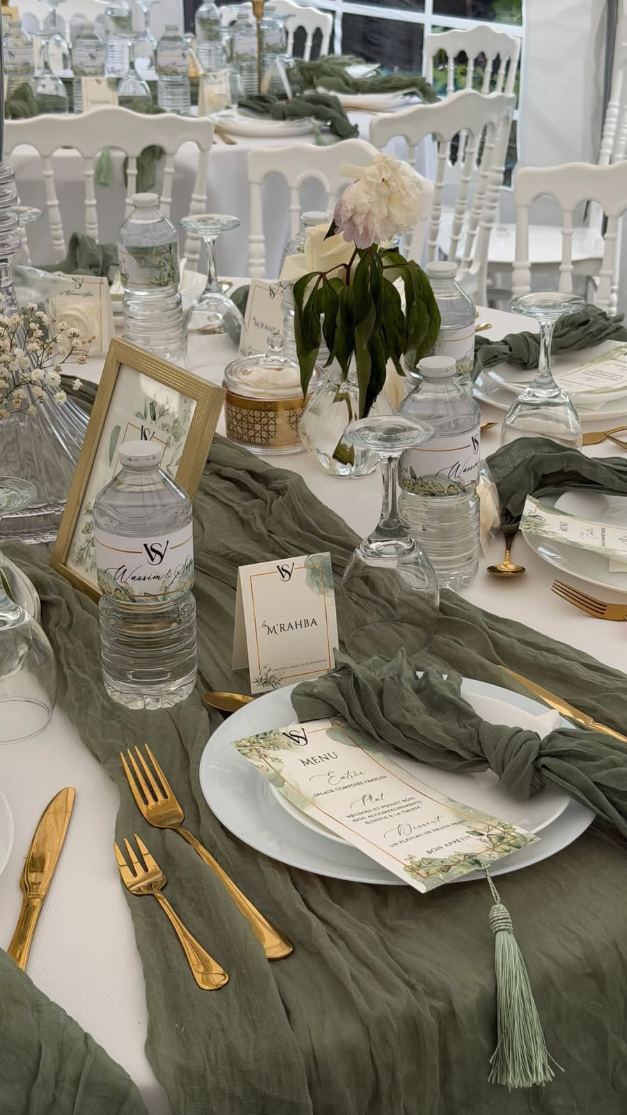 Table dressée sous chapiteau : chemin de table en gaze vert sauge, couverts dorés, menu calligraphié et carte M’rahba